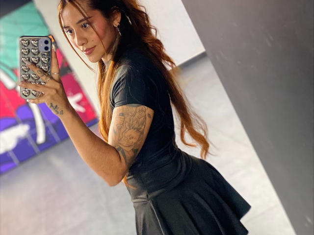 AlanaQuin with long brown hair in a black dress, showing off her tattooed arm and holding a phone with a pop socket in a mirror selfie. Her ass is prominent.