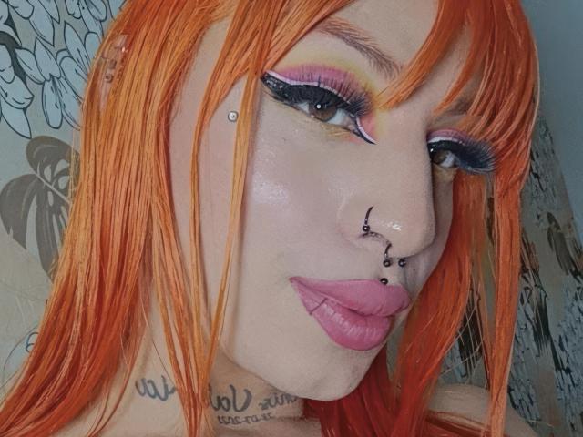 KeniaDolce with bright orange hair, pink lipstick, and multiple facial piercings, including a nose ring and septum stud, wearing dramatic blue and pink eyeshadow.
