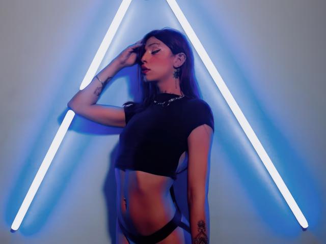 KeniaDolce, with her flat stomach and black crop top, stands against a blue neon triangle, hand in her brown hair, showing tattoos on her arm and hip.