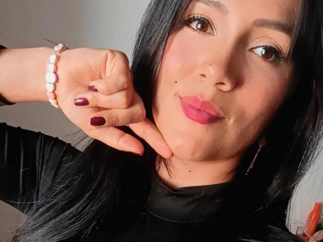 KathaxX has dark hair, pink lipstick, and a small beauty mark under her right eye. She's pointing at her chin with purple-painted nails, wearing a pink bead bracelet.