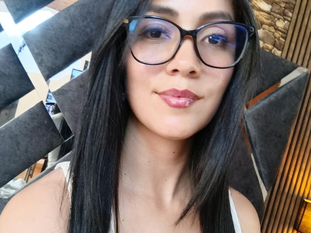 KathaxX with black glasses, pink lips, and long straight black hair, wearing a white tank top, smiling slightly. Her ass is not visible. Her tits are small and her pussy is not shown.