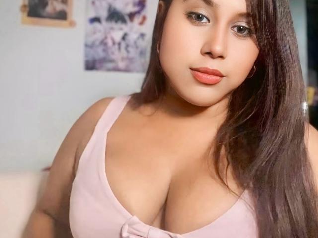CelestinneThomas with medium brown skin, long dark brown hair, pink tank top showing her big tits, pink lipstick, and a slight smile, looking at the camera.