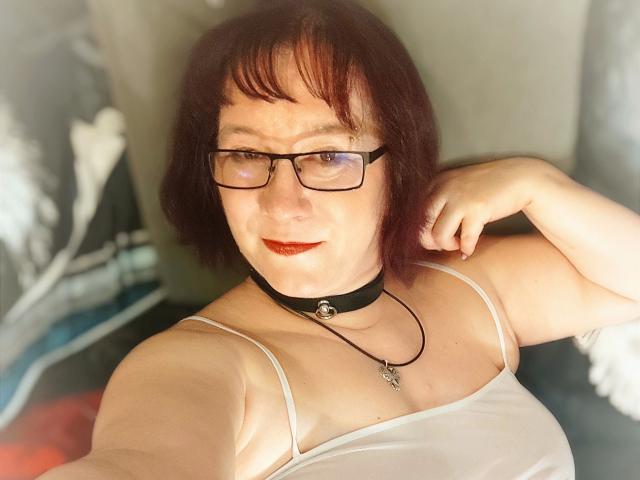 RotesTeufelchen-hot with short red hair, glasses, red lipstick, white tank top, black choker, and necklace, showing off her tits and a cute smile.