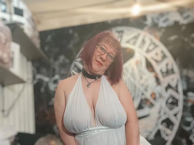 RotesTeufelchen-hot with purple hair, glasses, and a choker, wearing a gray halter dress that reveals her large, shiny tits. Soft lighting, slight smile, casual, indoors.