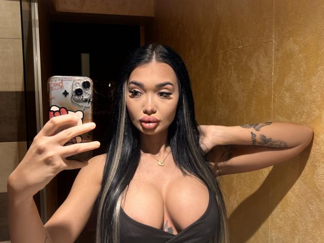 SashaFerrera tomando un selfie con cabello largo y negro, busto generoso y un tatuaje en su brazo derecho, vistiendo una blusa negra y sosteniendo un teléfono con una funda con forma de calavera..