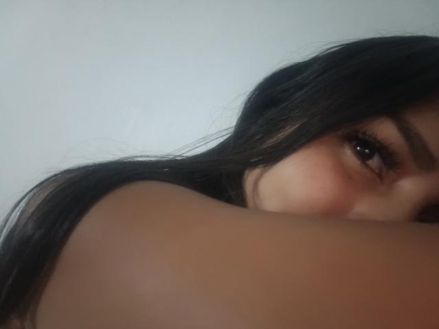 MelanySunny's face with dark hair, partially visible brown eye, and bare shoulder in a slightly blurred selfie. Her eye is slightly open, showing a subtle, sexy expression.