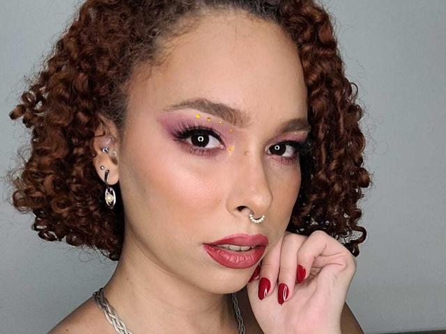 DominaMoon tiene el pelo castaño rizado, sombra de ojos morada, lápiz labial rojo, uñas rojas, un piercing en la nariz, un piercing en el tabique nasal, aretes y está tocándose la barbilla, con un fondo gris..