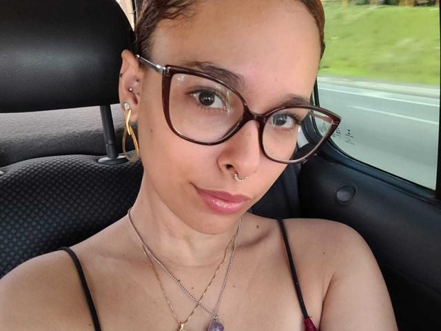 DominaMoon, avec des lunettes, un anneau de nez et des piercings aux oreilles, portant un top à bretelles fines, assise dans une voiture. Ses seins sont petits, et elle a un air mignon et confiant..