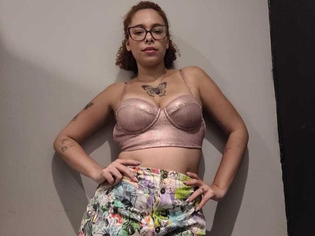 DominaMoon heeft krullend bruin haar, draagt een bril en een roze, glinsterende beha die haar borsten accentueert. Ze draagt ook kleurrijke broeken en heeft haar handen op haar heupen..