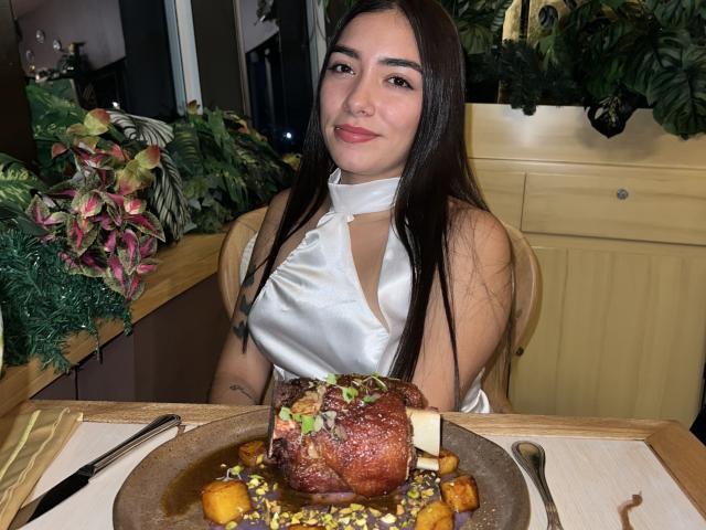 SharonKlart, avec de longs cheveux bruns, portant une robe en satin blanc à encolure dégagée, souriant devant un poulet rôti avec des oignons caramélisés et des petits pois sur une table en bois..