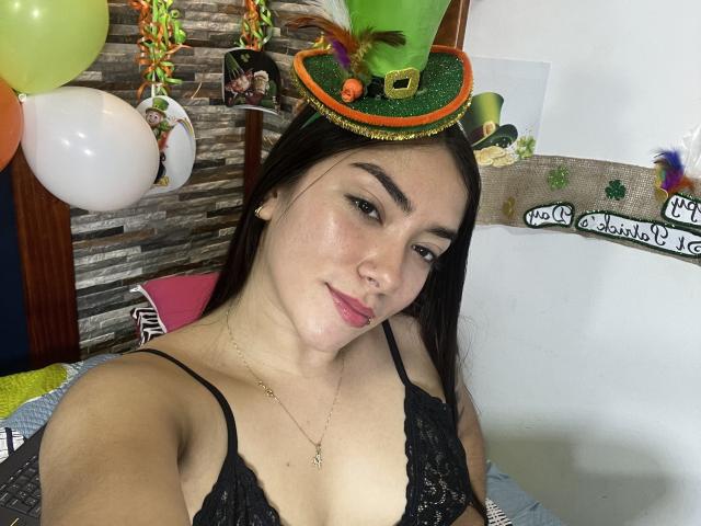 SharonKlart with black lace bra, green glitter hat, and colorful feathers, smiling, brown eyes, pink lipstick, long dark hair, light skin, small gold necklace, in a bedroom.