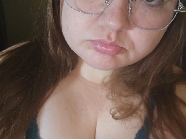SexxiJiji's face, showing glossy pink lips, round glasses, and brown hair, with a focus on her cleavage in a dark green top. Her boobs are partially visible.