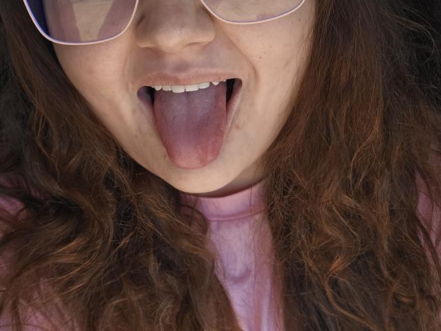 SexxiJiji's face with wavy brown hair, pink t-shirt, and glasses. She's sticking out her tongue, showing her teeth and a small freckle on her upper lip.
