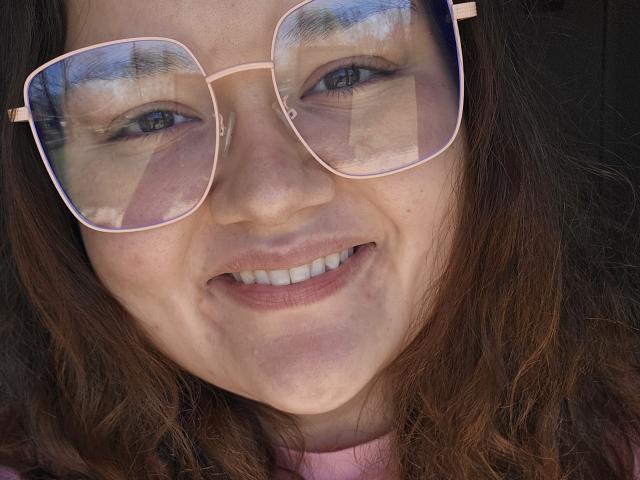 SexxiJiji has a cute smile with pink-tinted aviator sunglasses reflecting a building. She has wavy brown hair, blue eyes, and a light pink shirt. Her face is slightly flushed.
