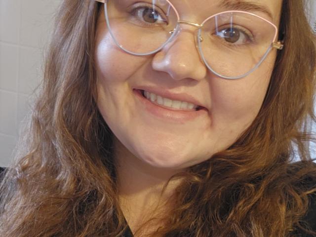 SexyJiji with wavy brown hair, wearing pink-rimmed glasses, smiling with slightly flushed cheeks, showing small, cute tits, and a cute, round ass, in a selfie.