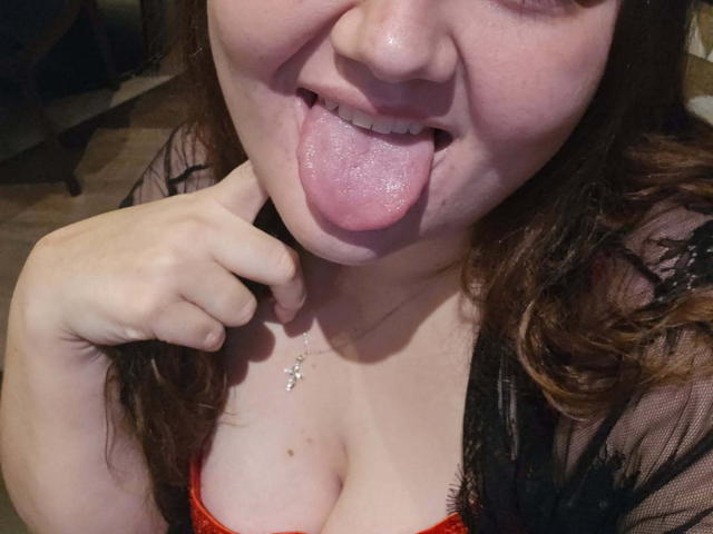 SexxiJiji has a chubby face, her tongue is out, and she's sticking her finger near her cleavage. She's wearing a red bra and a black lace robe.