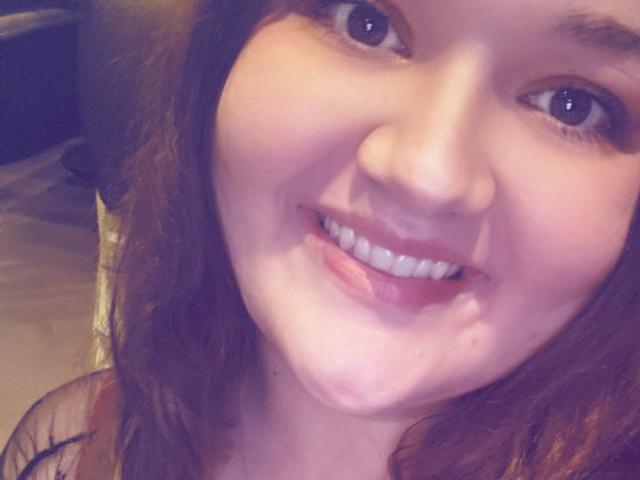SexxiJiji's smiling face with cum on her chin, brown wavy hair, blue eyes, and white teeth, wearing a black top, in a selfie.