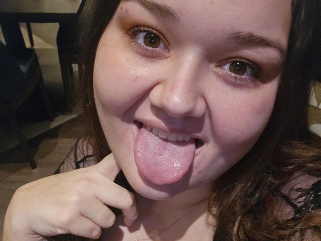 SexxiJiji with brown eyes, pink tongue out, slightly flushed face, and wavy brown hair, holding her chin. She has a small mole above her right eyebrow.