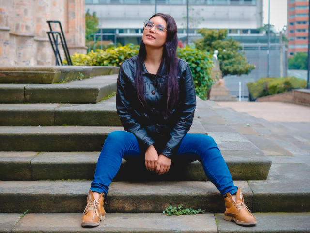 AriaLeenn with long dark purple hair, glasses, black leather jacket, blue jeans, and tan boots, sitting on stone steps with legs apart, hands on knees, smiling.
