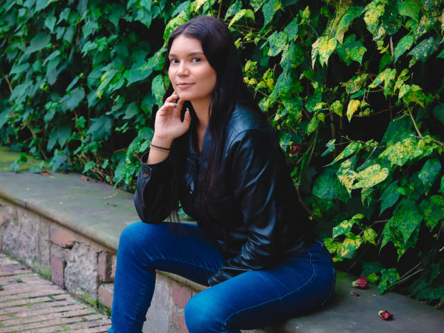 AriaLeenn with long black hair, black leather jacket, and blue jeans, sitting on a stone ledge, hand near her face, looking cute and confident.
