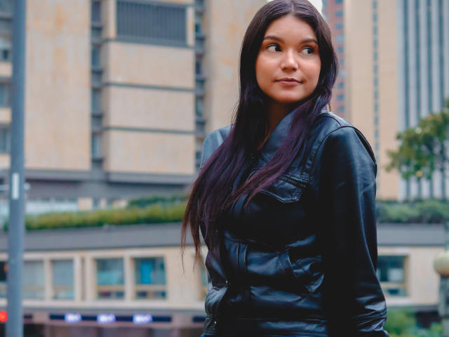 AriaLeenn with long dark purple hair, wearing a black leather jacket, looking to the side with a slight smile, standing in an urban setting, showing her tits subtly.