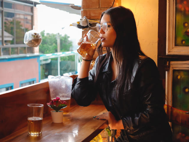AriaLeenn with long dark hair, glasses, black leather jacket, sipping beer, sitting at wooden table, potted plant, glass of beer, bird ornament, warm yellow light, casual vibe.