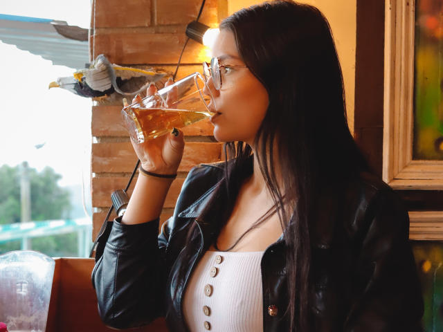AriaLeenn with long black hair, glasses, black leather jacket, white button-up, drinking from a glass, holding a bird, warm light, brown brick wall, casual urban setting.