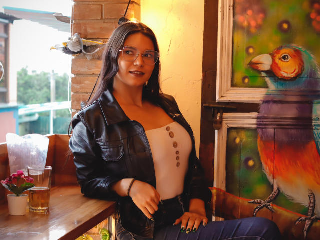 AriaLeenn with long dark hair, glasses, black leather jacket, white tank top showing tits, and blue jeans, sitting next to a colorful bird painting in a cozy cafe.