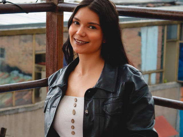 AriaLeenn, with dark brown hair and a smile, wears a black leather jacket over a white ribbed top with button details, standing near rusty metal bars.