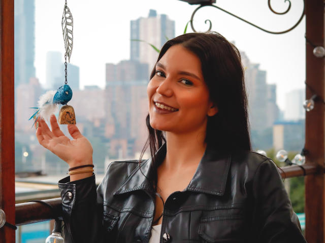 AriaLeenn with dark hair, smiling, holding a blue feather ornament, wearing a black leather jacket, showing her tits, in a wooden, outdoor setting, city skyline in the background.