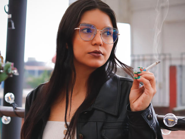 AriaLeenn with long black hair, wearing glasses, a black leather jacket, and a white top, smoking a cigarette with blue-green nails, looking serious.