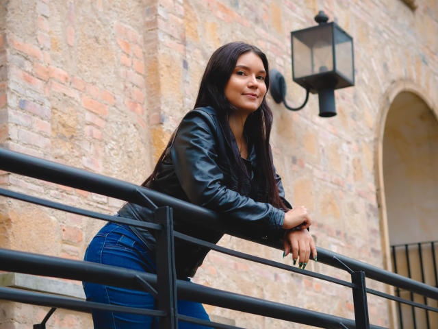 AriaLeenn with long black hair, leather jacket, and blue jeans, leaning on a black metal railing, smiling, hands resting on the rail, brick wall with lantern in background.