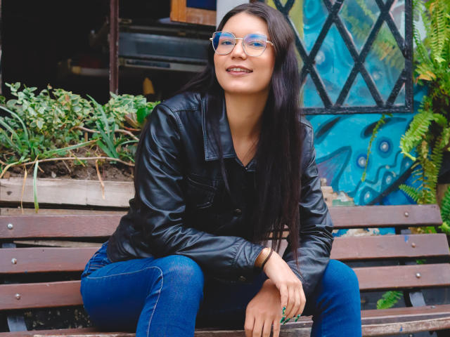 AriaLeenn with long black hair, wearing glasses, black leather jacket, and blue jeans, sitting on a wooden bench, smiling, hands on knee, green foliage in background.