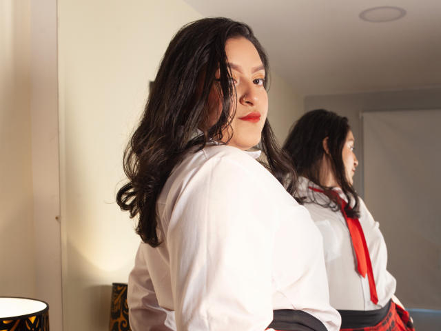 ShopiaRosse with long black hair, wearing a white shirt and red tie, looking over her shoulder, reflection in mirror, slightly open mouth, red lipstick, soft light, bedroom setting.