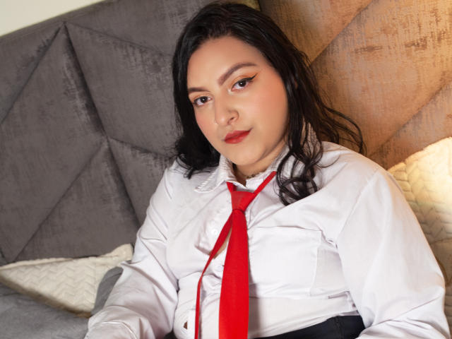 ShopiaRosse with dark hair, wearing a white shirt and red tie, sitting on a gray, tufted headboard, looking at the camera with a slight smile. Her tits are small.