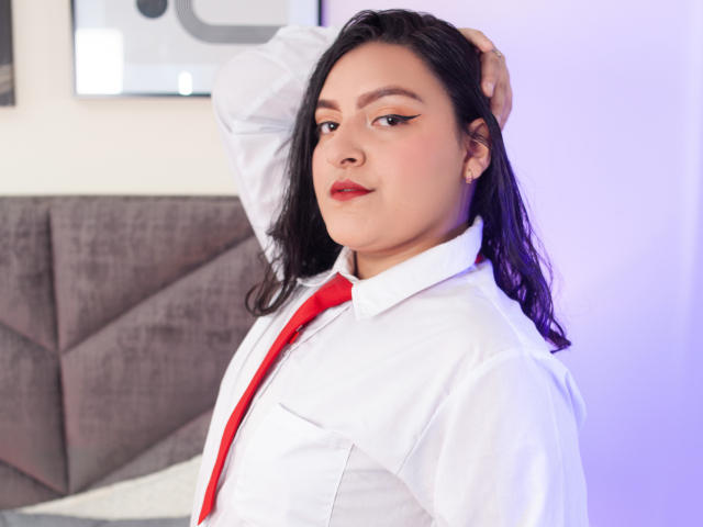 ShopiaRosse with black hair, white shirt, red tie, and red lipstick, looking confident, hand in hair, slight smile, soft pink lighting, and gray upholstered headboard in background.
