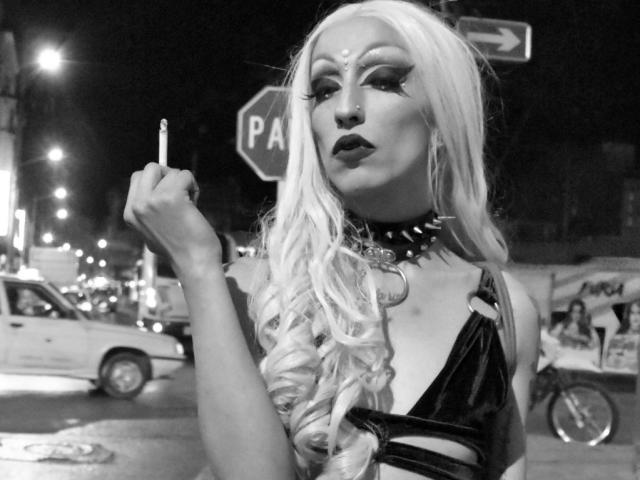 Fotografia in bianco e nero di FridaFendy con lunghi capelli biondo platino, trucco pesante, che tiene una sigaretta, indossa una collana con borchie e una top in pelle nera, mostra il seno, in piedi in una strada..