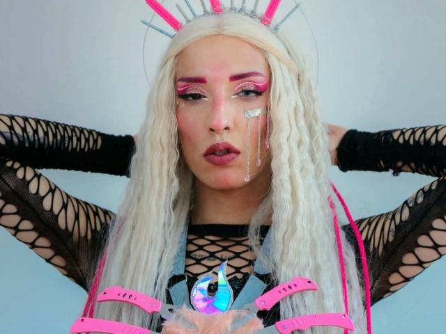 FridaFendy has platinum blonde dreads, pink eyeshadow, black fishnet sleeves, pink bunny ears, and a neon pink harness with a crystal pendant, looking serious with hands behind head.