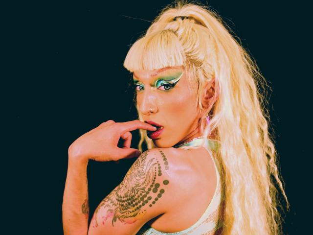 Blonde FridaFendy with green eye makeup, tongue slightly out, finger near mouth, tattooed arm, and long ponytail. She's looking back with sultry eyes, her tits barely visible.