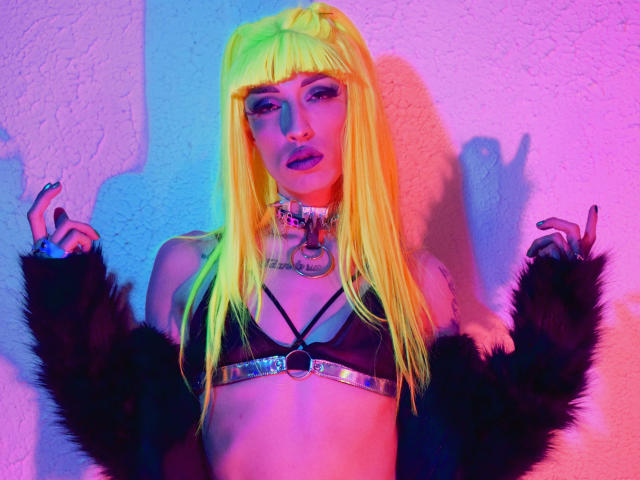 FridaFendy mit neon-gelbem Haar, einem schwarzen Top mit Trägern, einer schwarzen, fellbesetzten Jacke, einer metallenen Kette und pink-blauer Neonbeleuchtung, die eine doppelte Friedensgeste macht. Ihre Brüste sind sichtbar..