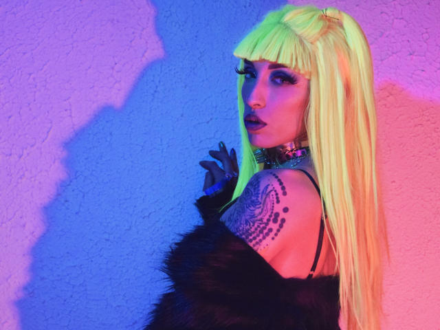 FridaFendy mit neon-grünem Haar, einem schwarzen Felloberteil, das von einer Schulter abrutscht, einem tätowierten Arm, dunklem Lippenstift und pink-blauer Beleuchtung. Ihre Brüste sind teilweise sichtbar, während sie über ihre Schulter schaut..