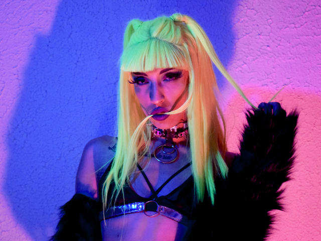 FridaFendy with neon blonde pigtails, black leather harness, purple lipstick, choker with ring, black furry jacket, holding hair, neon blue and pink lighting.