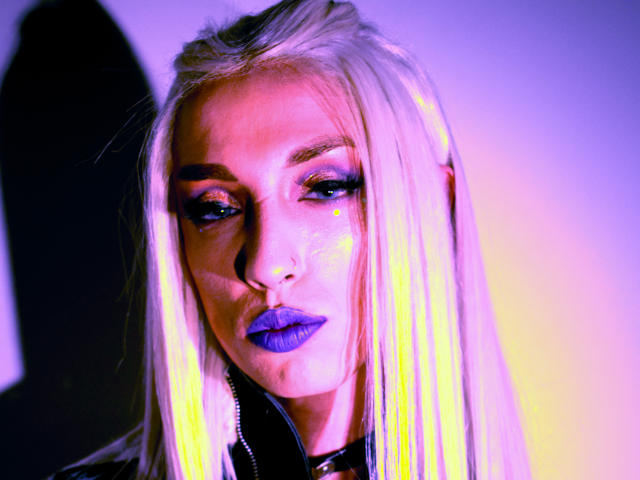 FridaFendy has long blonde hair, blue lipstick, and glittery eye makeup. Her face is lit with neon purple and yellow. She's looking down with a serious expression.