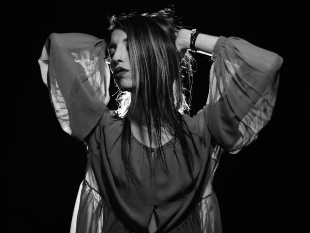 Fotografia in bianco e nero di FridaFendy con i capelli lunghi, le braccia alzate, che indossa una maglia trasparente con maniche a sbuffo, che guarda di lato, sfondo scuro, braccialetto visibile al polso..