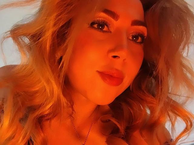 DouceKhrystine has wavy blonde hair, orange-tinted skin, full eyebrows, and red lipstick. She's looking at the camera with a slight smile, wearing a necklace.