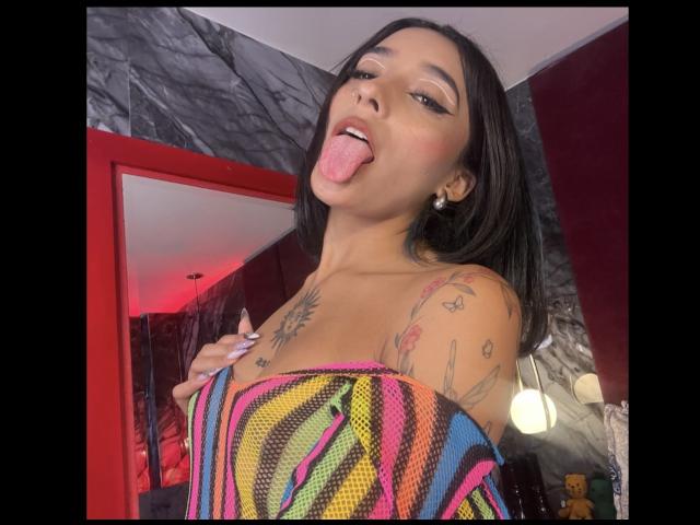 SalomeMille tem cabelo escuro, está de língua para fora, vestindo uma blusa colorida listrada com um ombro de fora, mostrando tatuagens no peito e no ombro, com uma expressão divertida..