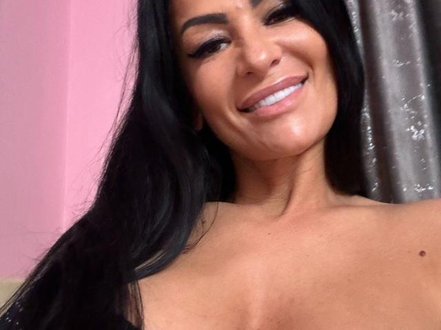 AndraD has long black hair, a smiling face, and is topless, showing her tits. She has a small mole on her right shoulder and is wearing white teeth.