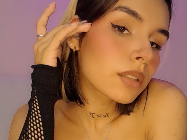 LenaBranch hat ein süßes Gesicht mit dunklen und blonden Haaren, einem schwarzen Netzhandschuh, einem Nasenring und einem Tattoo am Hals. Sie schaut mit einem leichten Lächeln in die Kamera..