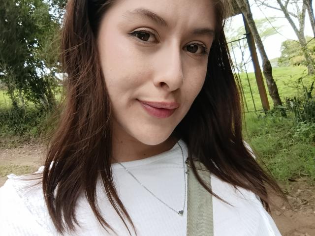 IsabellaPalacio has light brown skin, brown eyes, and wavy brown hair. She's wearing a white shirt with a green strap, and a silver necklace, smiling softly.