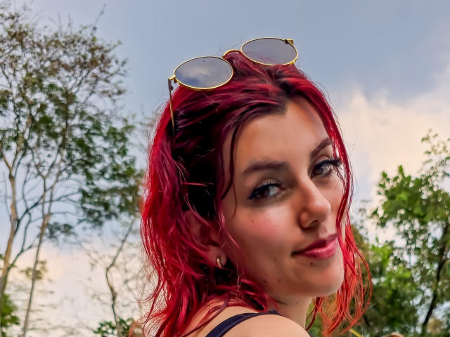 ZoeeMar met rood haar, zonnebril op haar hoofd, eyeliner, neusring, oorbellen, lachend, kijkt over haar schouder, buiten, draagt een zwarte top, met een boom en de lucht op de achtergrond..